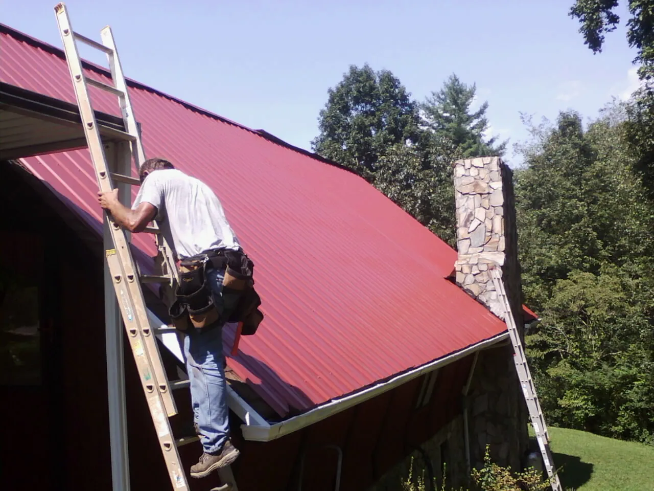 Roofer on a ladder beside a red metal roof and stone chimney — Soffit & Fascia Repair by Roofing Brothers in Opelousas